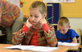 Girl pictured sitting at a table in a classroom with a boy sitting at another desk behind her