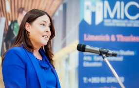 Dr Gwen Moore dressed in a blue suit pictured speaking into a microphone against a background that shows a picture of the MIC logo