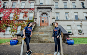 Two female students wearing MIC t-shirts and jeans lean on silver poles outside the steps leading up to a door on the Foundation Building at MIC