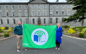 A man & woman pictured holding the Green Campus Flag on the grounds of MIC Thurles with the MIC Thurles building in the background