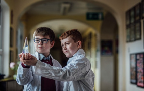 Two boys in science coats holding and looking at a beaker