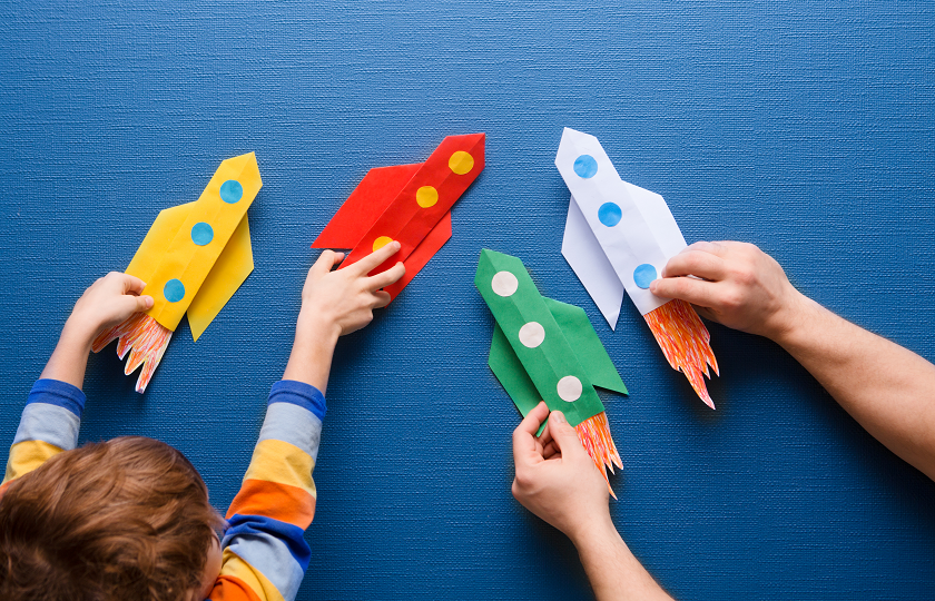 Child and adult playing with paper rockets against a blue backdrop 