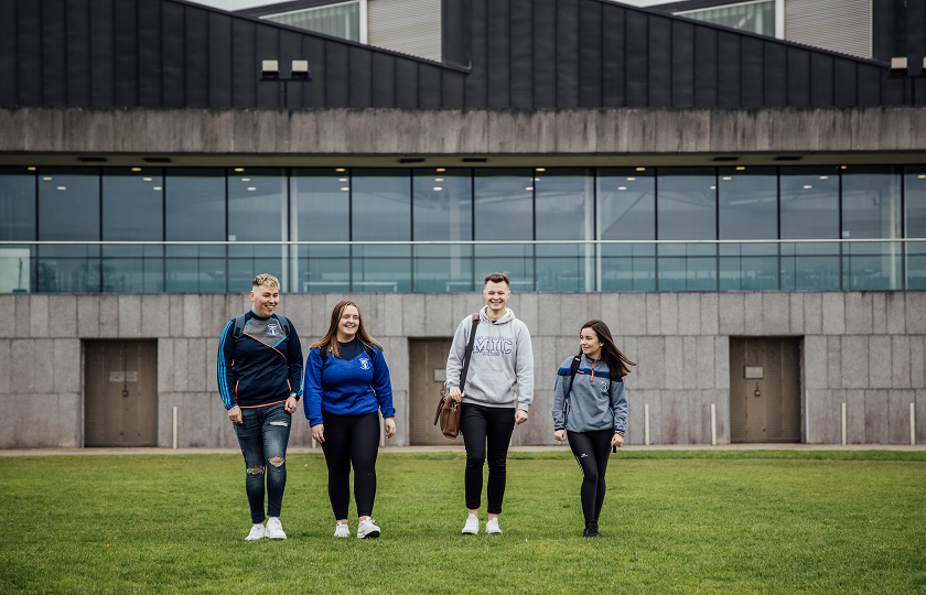 A group of four students are walking across a green field. There is a large grey building in the background.