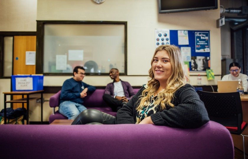 Female student with blonde hair sits on a purple couch looking over her shoulder with a smile at the viewer. There are a number of student sitting behind her in the background and are talking.