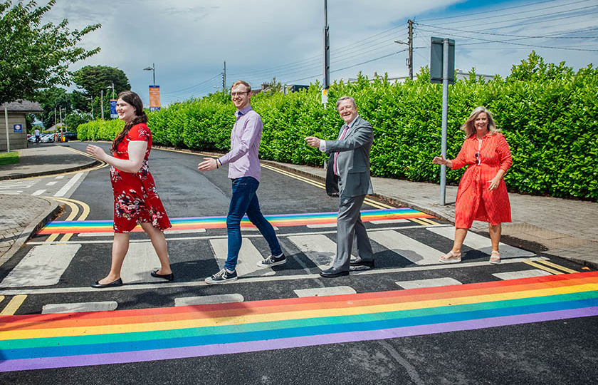 Four people walk single file across a pedestrian crossing with rainbow trim